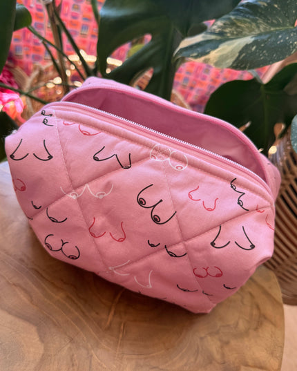 A pink quilted cosmetic pouch with a black and pink cat face pattern, resting on a wooden surface next to green houseplants.