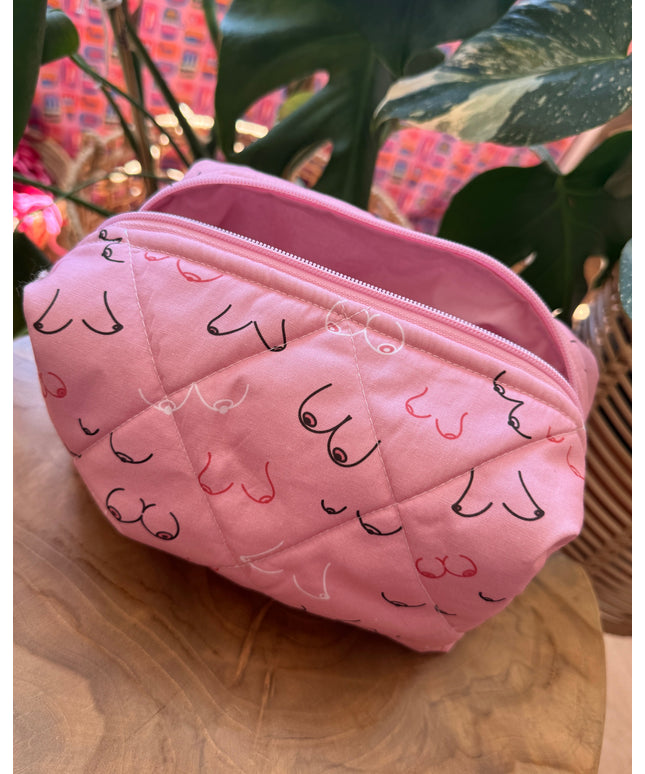 A pink quilted cosmetic pouch with a black and pink cat face pattern, resting on a wooden surface next to green houseplants.